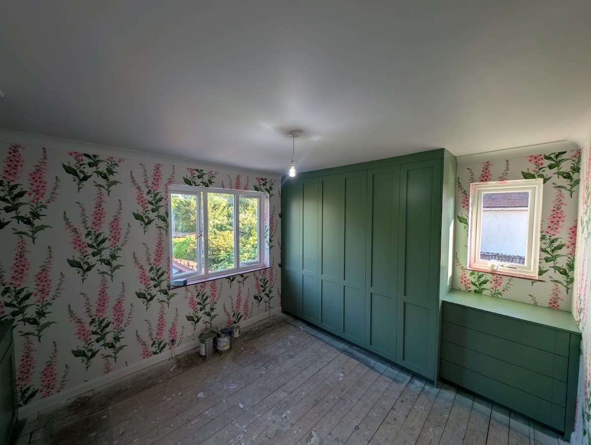 Wide view of a bedroom with sage green fitted wardrobes, a built-in window seat and pink foxglove-pattern wallpaper on the surrounding walls