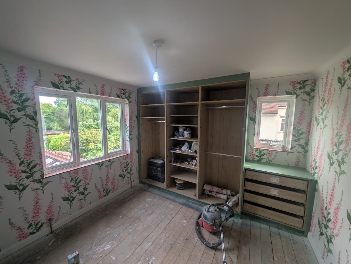A foxglove-pattern wallpapered bedroom with an open wardrobe carcass fitted against one wall and a green drawer unit under the window