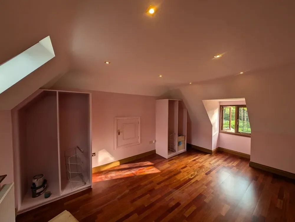 Finished loft bedroom with sloped ceilings, recessed lights and built-in wardrobe recesses, all in an even soft pink finish above a wooden floor