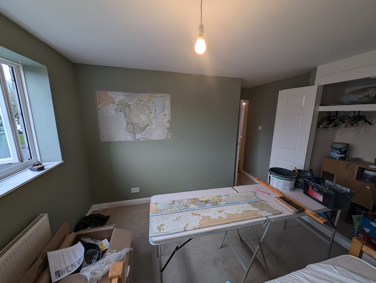 A bedroom mid-installation, with world map wallpaper panels laid out on a trestle table for cutting and one panel already mounted on the wall